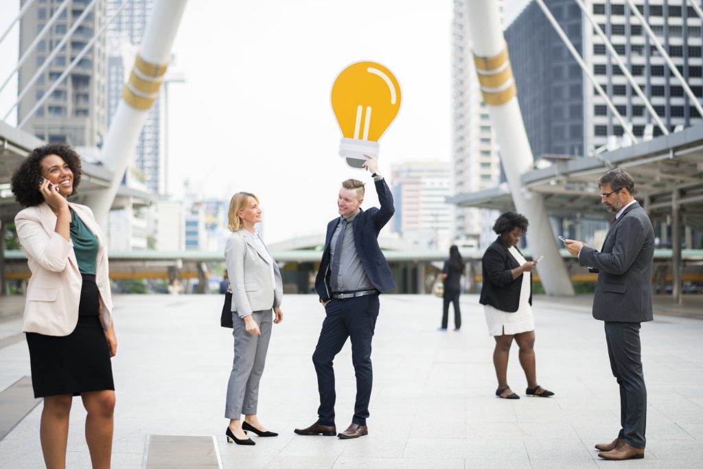 Five businesspeople in suits stand outdoors in a city. A man playfully holds a large light bulb cutout above his head, symbolising an idea or innovation